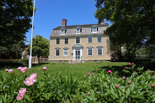 A historic yellow house with white trim, surrounded by green grass and pink flowers under a clear blue sky.