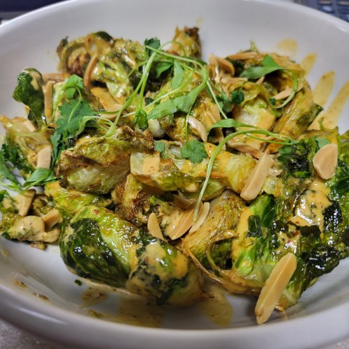 A bowl of roasted Brussels sprouts topped with herbs, sliced almonds, and a creamy sauce.