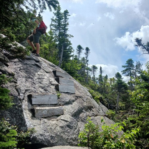 A hiker climbs a rocky outcrop surrounded by lush trees and a bright blue sky with scattered clouds.