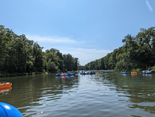 A serene river scene with colorful inflatable rafts floating among lush green trees under a clear blue sky.