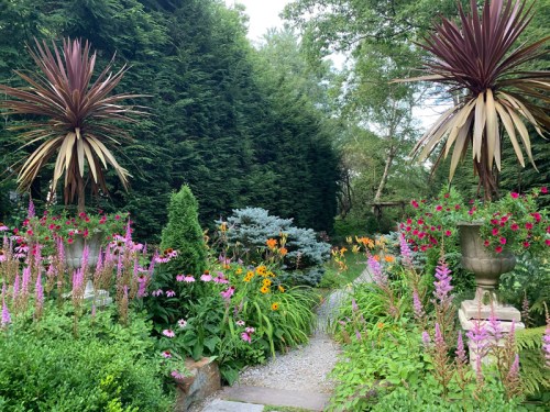 A lush garden path surrounded by vibrant flowers and tall plants, leading through greenery and decorative urns.