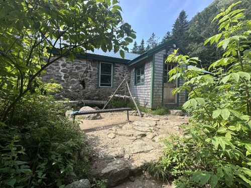 A rustic stone cabin surrounded by greenery and trees, with a swing set in the foreground.