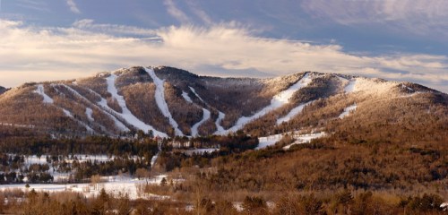 A panoramic view of snow-covered mountains with ski trails, surrounded by trees under a partly cloudy sky.