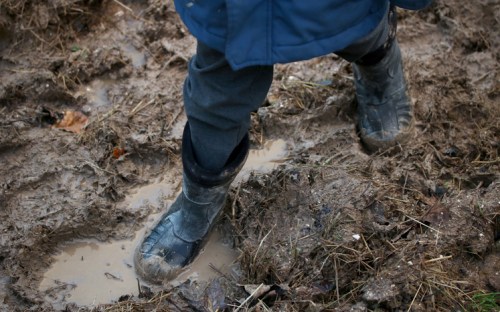 A person wearing black rubber boots stands in muddy ground with puddles and scattered leaves.