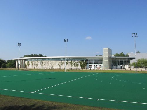 A modern sports facility with large windows, surrounded by a green field under a clear blue sky.