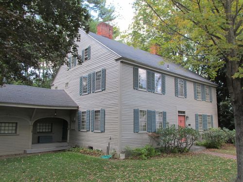 A two-story house with gray siding, green shutters, and a red door, surrounded by trees and a grassy yard.