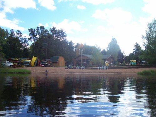 A serene lakeside view featuring a sandy beach, cabins, and colorful kayaks under a blue sky with scattered clouds.