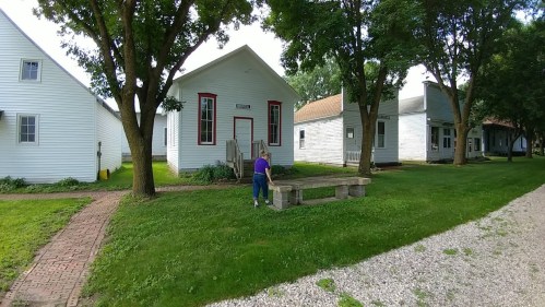 A person stands by a wooden bench in front of a white building surrounded by trees and other white structures.