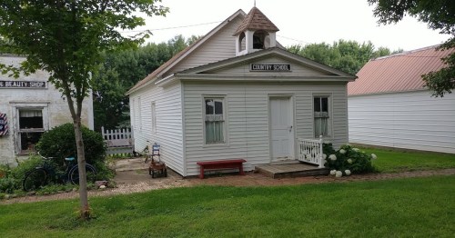 A quaint, white country schoolhouse with a porch, surrounded by greenery and a few bicycles nearby.