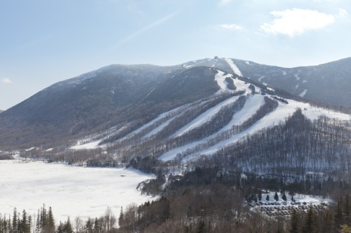 Snow-covered mountain with ski slopes, overlooking a frozen lake and trees under a clear blue sky.
