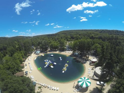 Aerial view of a sandy beach area with a circular pond, inflatable water toys, and green trees under a blue sky.