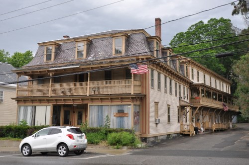 Historic building with a wooden porch, multiple stories, and an American flag, set against a cloudy sky.