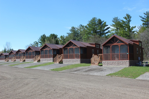 Row of rustic cabins with porches, set against a backdrop of trees and blue sky. Gravel path in the foreground.