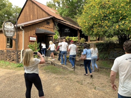 A group of people walking towards a rustic building with a sign for "Tasting Sales" surrounded by greenery.