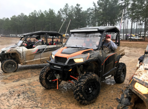 Two off-road vehicles in a muddy area, surrounded by trees, with people inside one vehicle and a person waving from another.