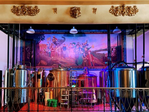 A colorful mural depicting people in traditional attire behind shiny brewing equipment in a vibrant setting.