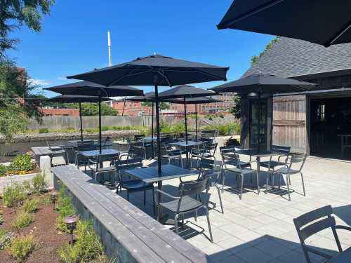 Outdoor dining area with black umbrellas over tables, surrounded by greenery and a clear blue sky.