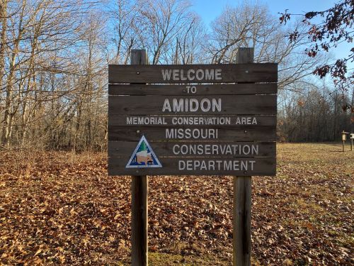 Wooden sign reading "Welcome to Amidon Memorial Conservation Area, Missouri Conservation Department," surrounded by trees and leaves.