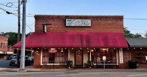 A brick building with a maroon awning, featuring a sign for "Big Zach's Place" and outdoor lighting.