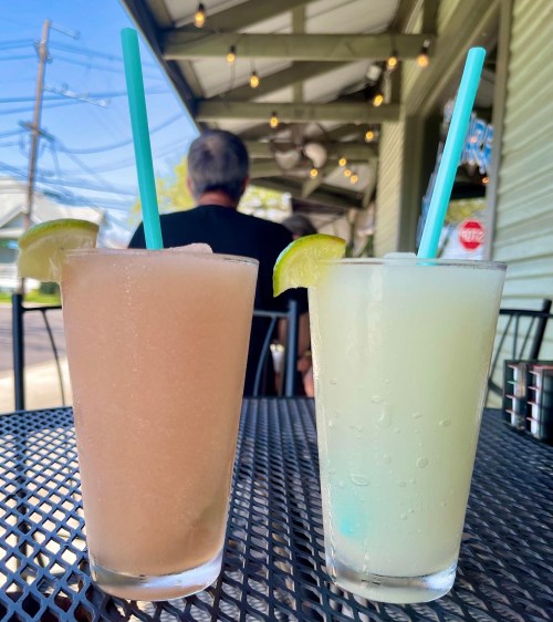 Two colorful drinks with lime wedges sit on a table, with a blurred background of people and outdoor seating.