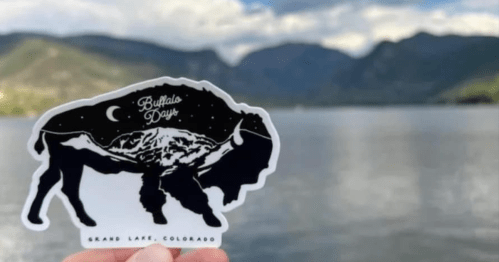 A hand holds a black and white sticker of a bison against a scenic lake and mountain backdrop in Grand Lake, Colorado.