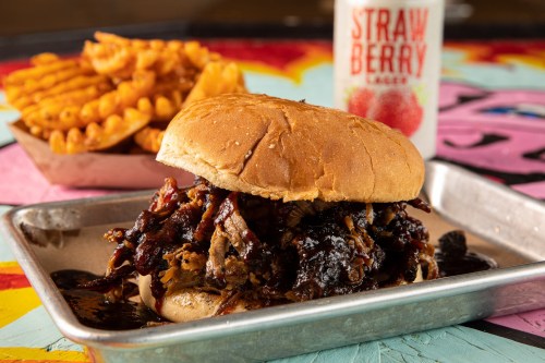 A close-up of a pulled pork sandwich with barbecue sauce, served with crinkle-cut fries and a strawberry drink in the background.