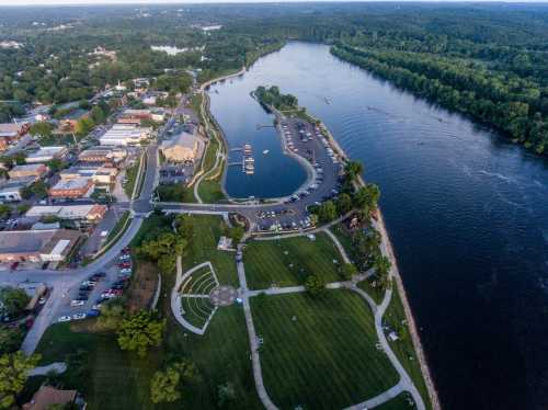 Aerial view of a riverside park with green spaces, a marina, and nearby town buildings along the water's edge.