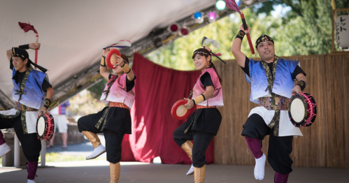A group of performers in colorful costumes dance on stage, playing traditional drums at a cultural festival.