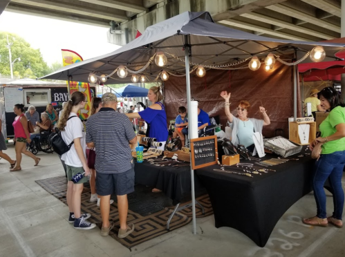 A bustling market scene with vendors, shoppers, and a tent displaying handmade jewelry under string lights.