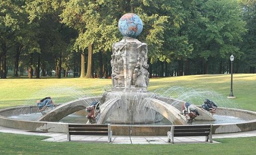 A decorative fountain featuring a globe atop a sculpted base, surrounded by water jets in a green park setting.