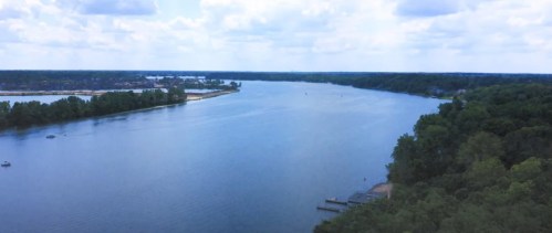 A serene river flows through lush greenery under a cloudy sky, with boats and docks visible along the shore.
