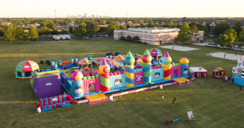 Aerial view of a colorful inflatable bounce house park set up on a grassy field with a city skyline in the background.