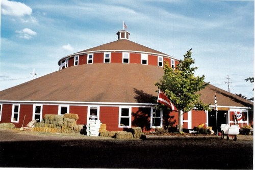 A round red barn with a circular roof, surrounded by hay bales and flags, under a blue sky with clouds.