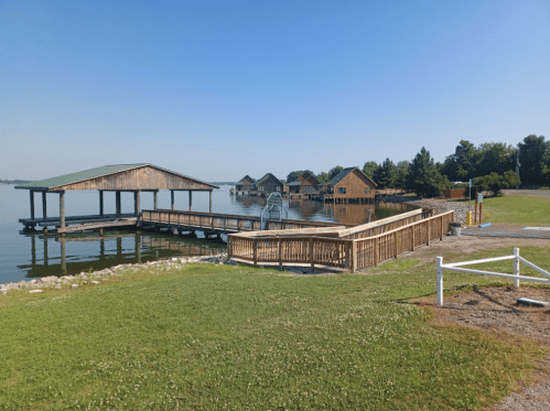 A wooden dock extends over calm water, with houses visible in the background and a clear blue sky above.