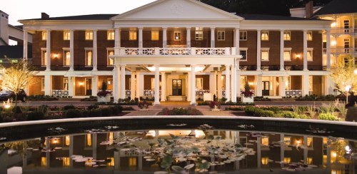 Elegant brick hotel with white columns, reflecting in a pond with lily pads, illuminated at dusk.