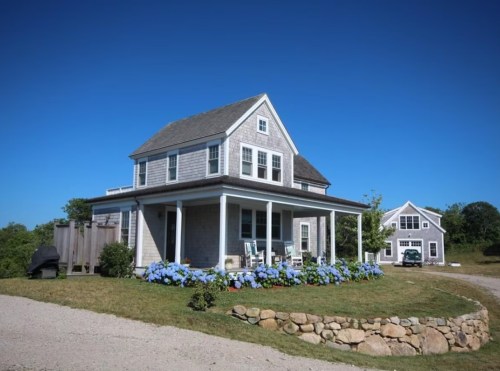 A charming two-story house with a porch, surrounded by blue hydrangeas and a clear blue sky.