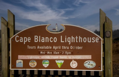 Sign for Cape Blanco Lighthouse, detailing tour hours and information, with the lighthouse visible in the background.
