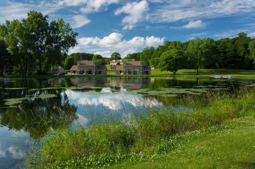 A serene lake reflects a blue sky and clouds, surrounded by lush greenery and a modern building in the background.