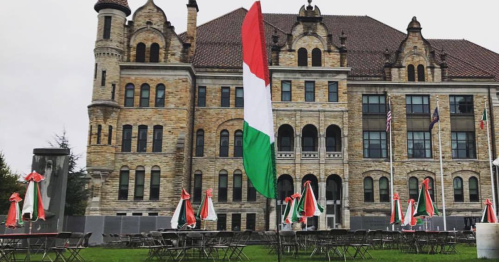 A green, white, and red flag stands in front of a historic stone building with outdoor seating and umbrellas.