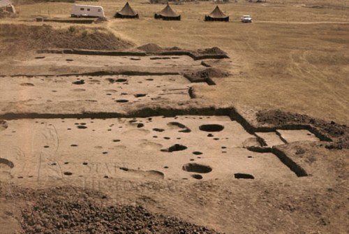 Aerial view of an archaeological site with excavation pits, tents, and a trailer in a grassy landscape.