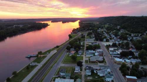 Aerial view of a river at sunset, reflecting vibrant colors, with a small town and green hills in the background.