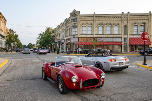 Two classic cars, a red convertible and a silver sports car, drive through a charming downtown street with historic buildings.
