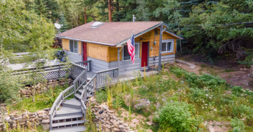 A cozy cabin surrounded by trees, featuring a wooden deck, stone pathway, and an American flag hanging by the door.