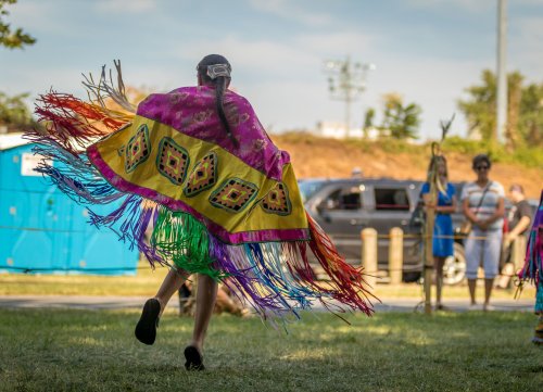 A dancer in a colorful fringed shawl performs at an outdoor event, with spectators in the background.
