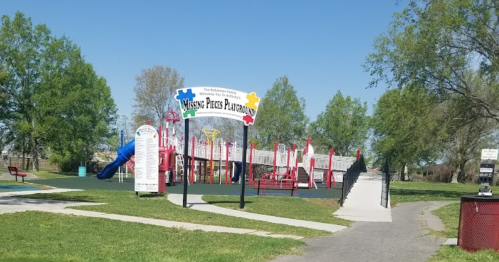 Colorful playground with slides and climbing structures, surrounded by trees and a walking path. Bright sunny day.