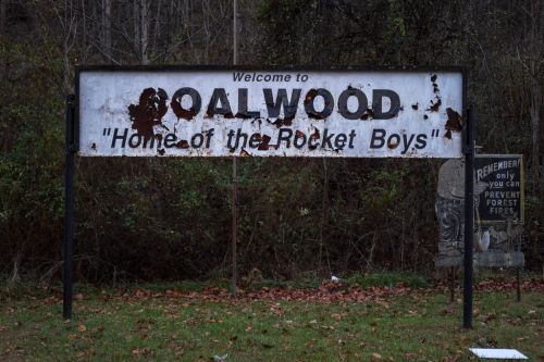 Weathered sign reading "Welcome to Coalwood, Home of the Rocket Boys," surrounded by trees and fallen leaves.