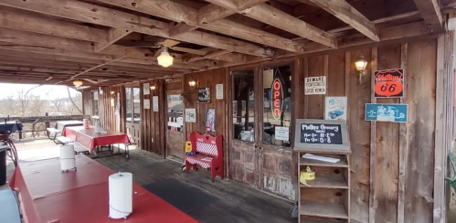 A rustic restaurant entrance with wooden walls, red tables, and various signs, including an "Open" sign and Route 66 memorabilia.