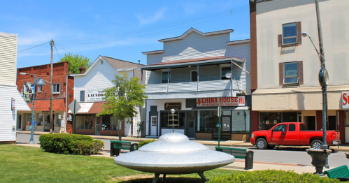 A small town street featuring a UFO sculpture, shops, and a red truck parked nearby under a clear blue sky.