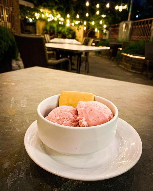 A bowl of pink ice cream with a cookie, set on a table outdoors with string lights in the background.
