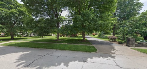 A peaceful neighborhood scene featuring green trees and a wide, paved road intersecting with a grassy area.